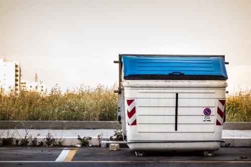 A large, white industrial waste skip positioned on an asphalt surface in an outdoor setting, with a blue tilting lid on top. The skip features red and white reflective chevron markings on its sides and a small circular no-parking symbol. It is situated adjacent to a raised curb and a road, with grass, wildflowers, and some tall plants in the background. The scene is illuminated by natural daylight, with a distant view of a horizon line and faint outlines of buildings or structures. This image is relevant to rubbish and waste collection services provided by Garden Clearance Swiss Cottage in the local area, highlighting the type of larger waste containers used for rubbish removal in urban environments.