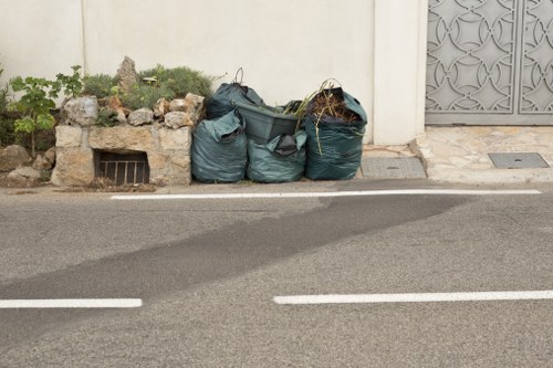 Three large, dark green plastic rubbish bags filled with garden waste and debris are placed on the pavement in front of a white wall with a small garden area containing plants and rocks to the left. A metal grate is partially visible beneath the planting area, and the bags are positioned beside a drain cover on the sidewalk. To the right, a decorative grey metal gate and a section of pavement are visible, indicating a residential setting near Swiss Cottage. The image captures a typical scene of garden clearance waste awaiting removal, with the rubbish bags aligned along the curbside, ready for collection by a local waste management service such as Garden Clearance Swiss Cottage. The street surface is asphalt with white road markings, and the lighting suggests daytime conditions.