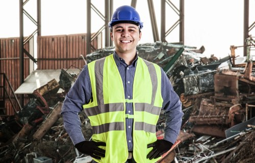 A male worker wearing a blue safety helmet, a high-visibility yellow vest over a long-sleeved shirt, and black gloves, standing inside a warehouse with large metal framework. Behind him is a large pile of assorted rubbish materials, including rusted metal parts and broken wooden structures, indicative of waste or debris from a demolition or clearance site. The lighting is natural, filtering through the warehouse's open panels, illuminating the scene clearly. The environment suggests a professional setting related to waste management or rubbish removal services, consistent with the offerings of Garden Clearance Swiss Cottage, which specializes in clearing rubbish from gardens and properties in the north London area, including Swiss Cottage postcode regions.
